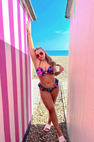 Woman in the butterfly bikini standing by a pink beach hut with a beach and ocean view.